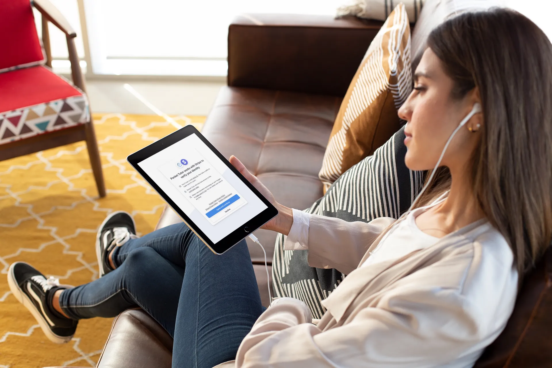 A woman sitting on a couch, holding an iPad, looking at a screen that says Pocket Tutor works with Stripe to verify your identity.